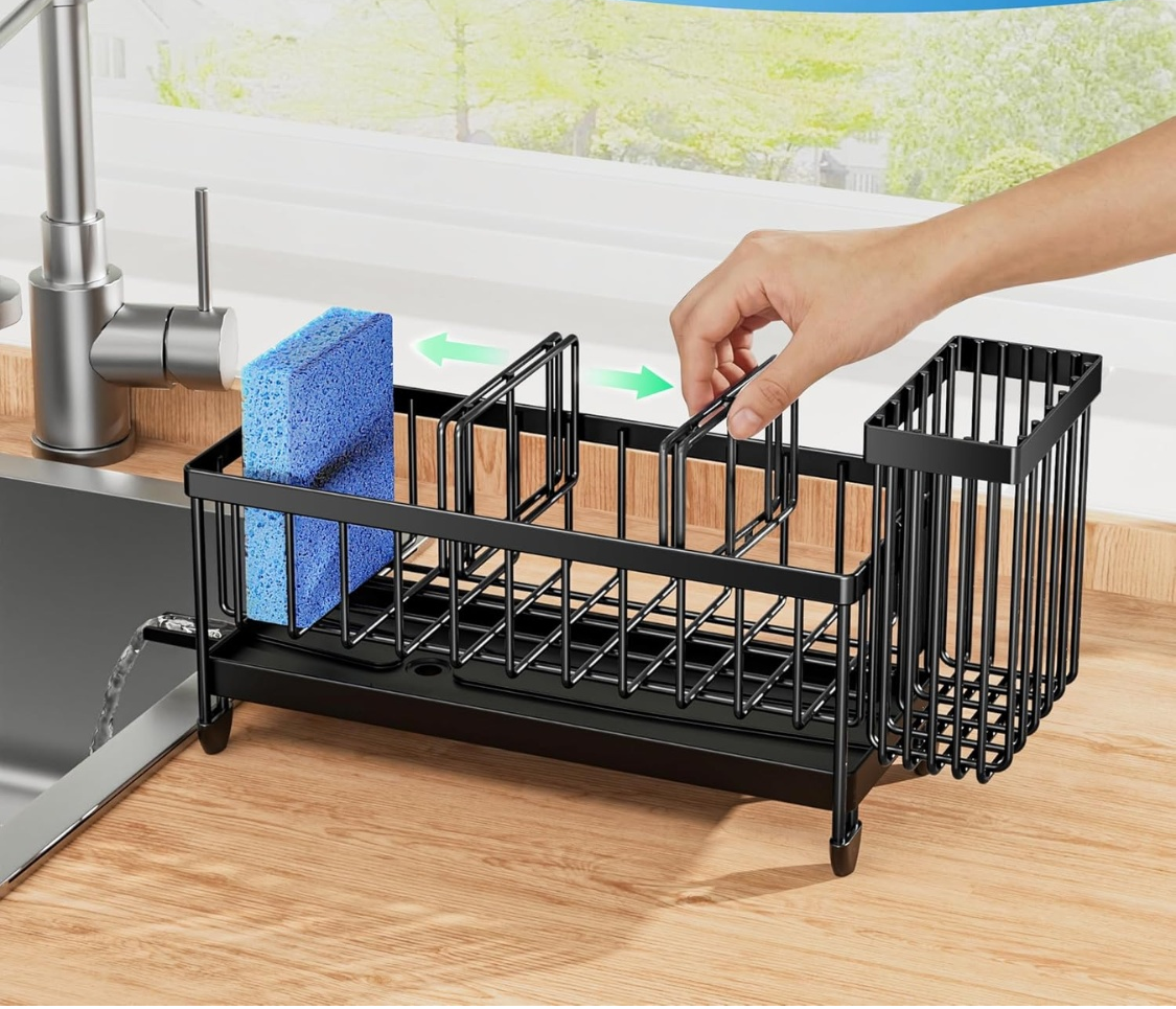 A black metal kitchen sink dish rack with a hand placing a blue sponge into one of its compartments, set on a wooden countertop next to a sink and faucet.